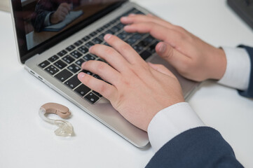 Close-up of a man's hands on a laptop keyboard. Hearing aid on the desktop.