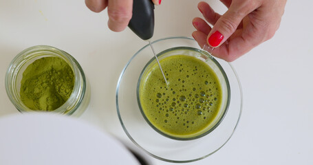 Woman is mixing matcha powder with hot water in glass cup by electric whisk 