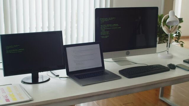 Medium close up of desk of programmer with computers with launched codes in testing mode on screens in office