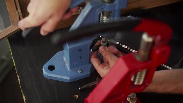 Close up view of professional leather craftsman attaches pin to zipper puller of handbag with hand press for installing metal fittings. Man with tattoo makes purse with special tool in workshop