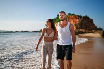Romantic couple walks on beach at sunset, holding hands. Newlyweds on honeymoon stroll seaside, barefoot in sand. Young man, woman enjoy sunset, love, ocean waves. Leisure, travel, holiday concept. © artiemedvedev