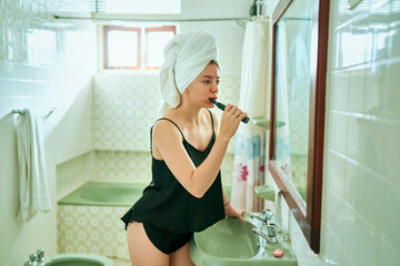 Young woman in bathroom brushing teeth, wearing towel on head and black underwear. Female practices...
