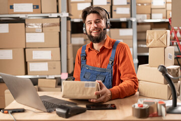 Caucasian male warehouse operator delivering goods dressed in blue overalls sits at a table holding a cardboard box in his hands, smiles and looks at the camera.