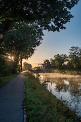 In this serene scene, the morning sun peeks through the trees along a tranquil waterway, casting a...