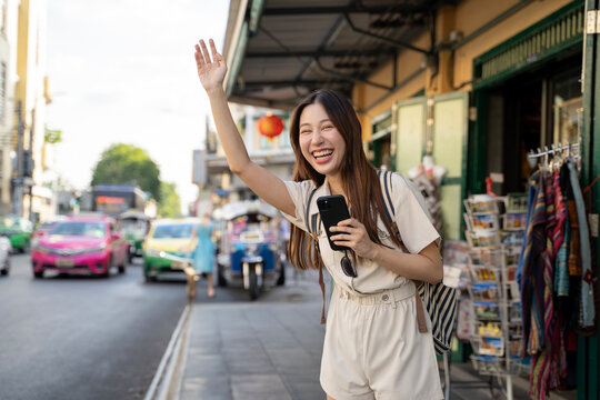 Young asian woman hailing a taxi in bustling Bangkok street, Thailand - Authentic urban travel moment capturing vibrant city life and local culture