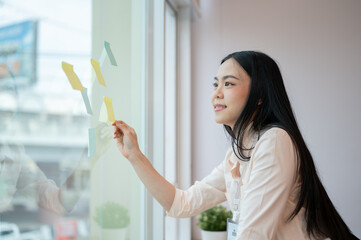 A beautiful Asian businesswoman attaching or looking at a sticky note on a glass wall.