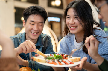 Group of cheerful and joyful young Asian friends enjoying eating food together at a restaurant. - obrazy, fototapety, plakaty