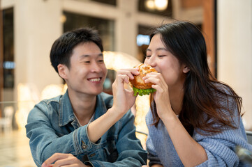 Cheerful young Asian couple enjoys eating a yummy burger on a weekend date at a restaurant.