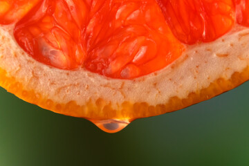 Red grapefruit juice or essential oil dripping from a fresh slice on a blurry green background, macro photography. Fruit background