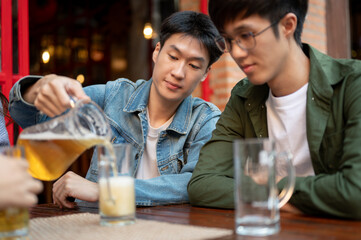 A happy Asian man is pouring beer for his friends while enjoying hanging out at a restaurant.