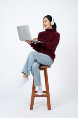 Young positive Asian woman in casual clothes wearing headphones and using her laptop on a chair.