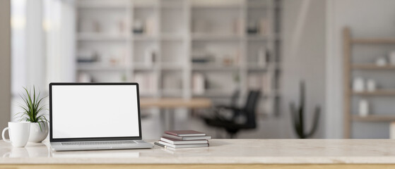 A white-screen laptop computer mockup, books, a mug, and a potted plant on a desk in a modern office