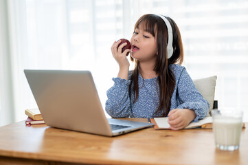 Young cute and happy Asian girl in headphones is eating an apple while studying online at home.