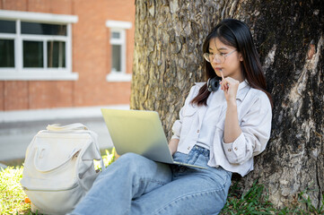 A positive Asian female student is using her laptop computer under the tree in a campus park.