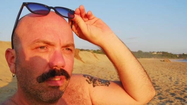 caucasian bald shaved head male with moustache and sunglasses at beach during sunset