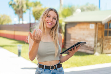 Naklejka na ściany i meble Young blonde woman holding a tablet at outdoors inviting to come with hand. Happy that you came