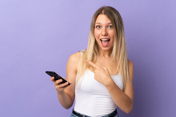 Young blonde woman using mobile phone isolated on purple background pointing finger to the side