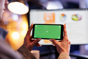 Businesswoman holding smartphone with green screen chroma key mock up display, working late at night in startup office. Executive manager analyzing marketing graphs report. Business concept