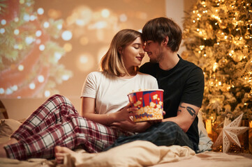 With popcorn. Beautiful young couple indoors, celebrating new year against projector