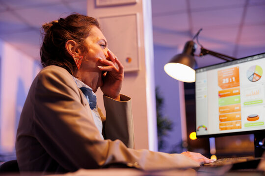 Exhausted manager yawning while analyzing financial graphs report on computer, working overhours at comany investment strategy in startup office. Tired sleepy entrepreneur typing profit progress
