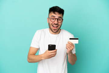 Young caucasian man isolated on blue background buying with the mobile and holding a credit card with surprised expression