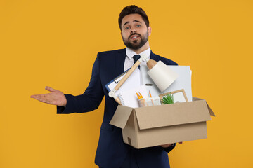 Confused unemployed man with box of personal office belongings on orange background