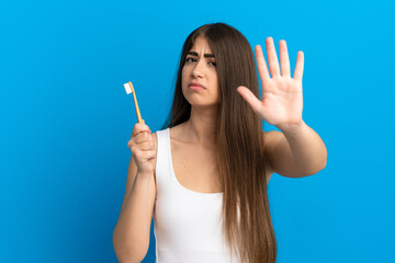 Young caucasian woman brushing teeth isolated on blue background making stop gesture and disappointed