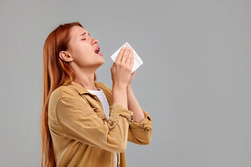 Suffering from allergy. Young woman with tissue sneezing on grey background, space for text