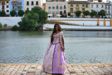 Beautiful latin woman with long curly hair wearing a 15th century dress is standing by the banks of the river guadalquivir in seville in spain. In the background you can see the other side of the city