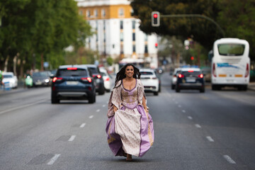 Beautiful Latin woman with long curly hair dressed in a 15th century dress running between cars on...