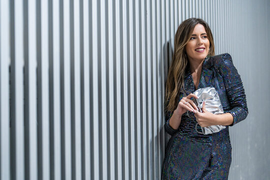 Smiling woman holding clutch bag and standing near striped wall
