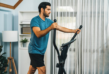Athletic and sporty man running on elliptical running machine during home body workout exercise session for fit physique and healthy sport lifestyle at home. Gaiety home exercise workout training. - obrazy, fototapety, plakaty