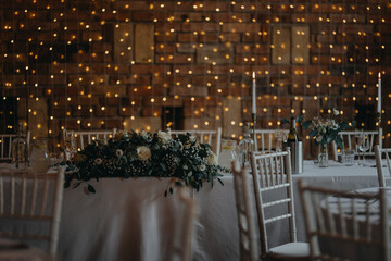 Elegant, romantic table setting for indoor wedding reception with fairy lights on wall behind. Luxury wedding table decoration, special event table set up with fresh flowers arragements.
