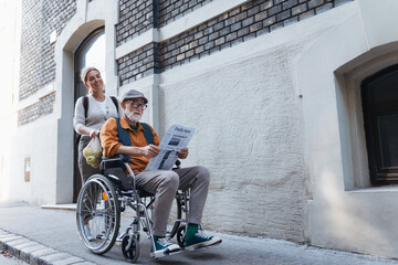 Granddaughter pushing senior man in wheelchair on street. Buying newspaper in newsstand. Female...