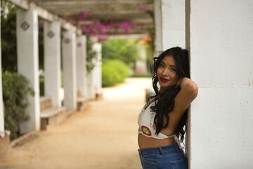 South American woman, young, beautiful, brunette, with crochet top and jeans leaning on a column, with a tender and dreamy look. Concept of beauty, fashion, trend, ethnicity, diversity.