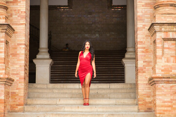 Fototapeta na wymiar South American woman, young, beautiful, brunette, with an elegant red dress, posing as a model in the square of Spain in Seville. Concept of beauty, fashion, trend, ethnicity, diversity, elegance.