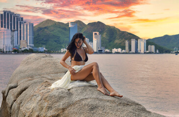 Young ukrainian woman in bikini and white dress sitting on a stone at the Nha Trang beach with...