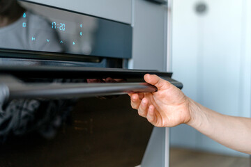 Selective focus on woman hand opening glass door of electric oven in modern kitchen.