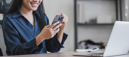 Happy Excited Asian young entrepreneur business woman using phone and laptop sitting on a desk at home workplace,