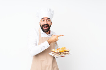 Young man holding muffin cake over isolated white background surprised and pointing side