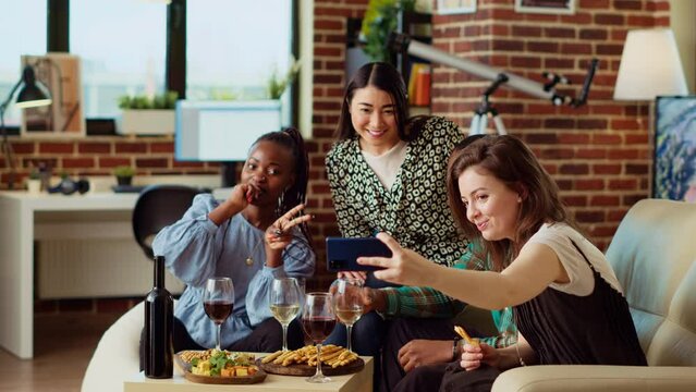 Multiracial friends taking group pictures with smartphone at apartment gathering. Cheerful people in living room posing for photo, making gestures and drinking alcoholic beverages
