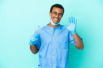 African American dentist holding tools over isolated blue background saluting with hand with happy expression
