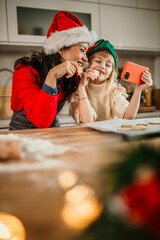 A heartwarming image of a mother and her daughter using a phone and tasting Christmas cookies in a festive kitchen