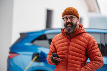 Man charging electric car during cold snowy day, using electric vehicle charging app, checking...