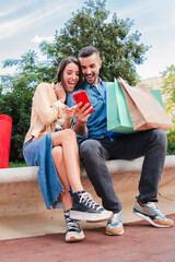 Vertical. Surprised couple using a cellphone to find sales, shopping online sitting with bags. Man...