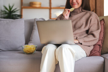 Closeup image of a young woman eating potato chips while watching on laptop computer at home