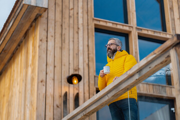 Man standing on cabins patio, drinking hot tea, coffee and enjoying beautiful winter day. Handsome man spending relaxing, stress-free winter weekend in cabin in mountains, enjoying alone time.