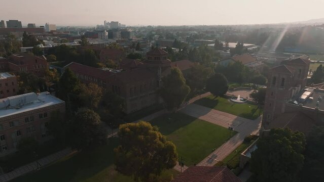 Aerial view of UCLA campus with Royce Hall center stage, Romanesque architecture, green spaces, and Westwood's urban backdrop in golden morning - afternoon light.