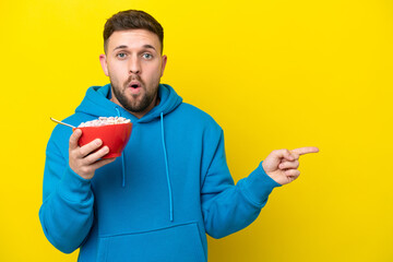 Young caucasian man holding a bowl of cereals isolated on yellow background surprised and pointing...
