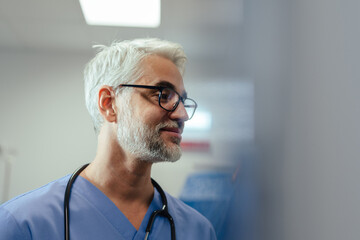 Portrait of confident mature doctor in hospital room. Handsome doctor with gray hair wearing medical scrubs and stethoscope around neck standing in modern private clinic, talking with patient, nurse.